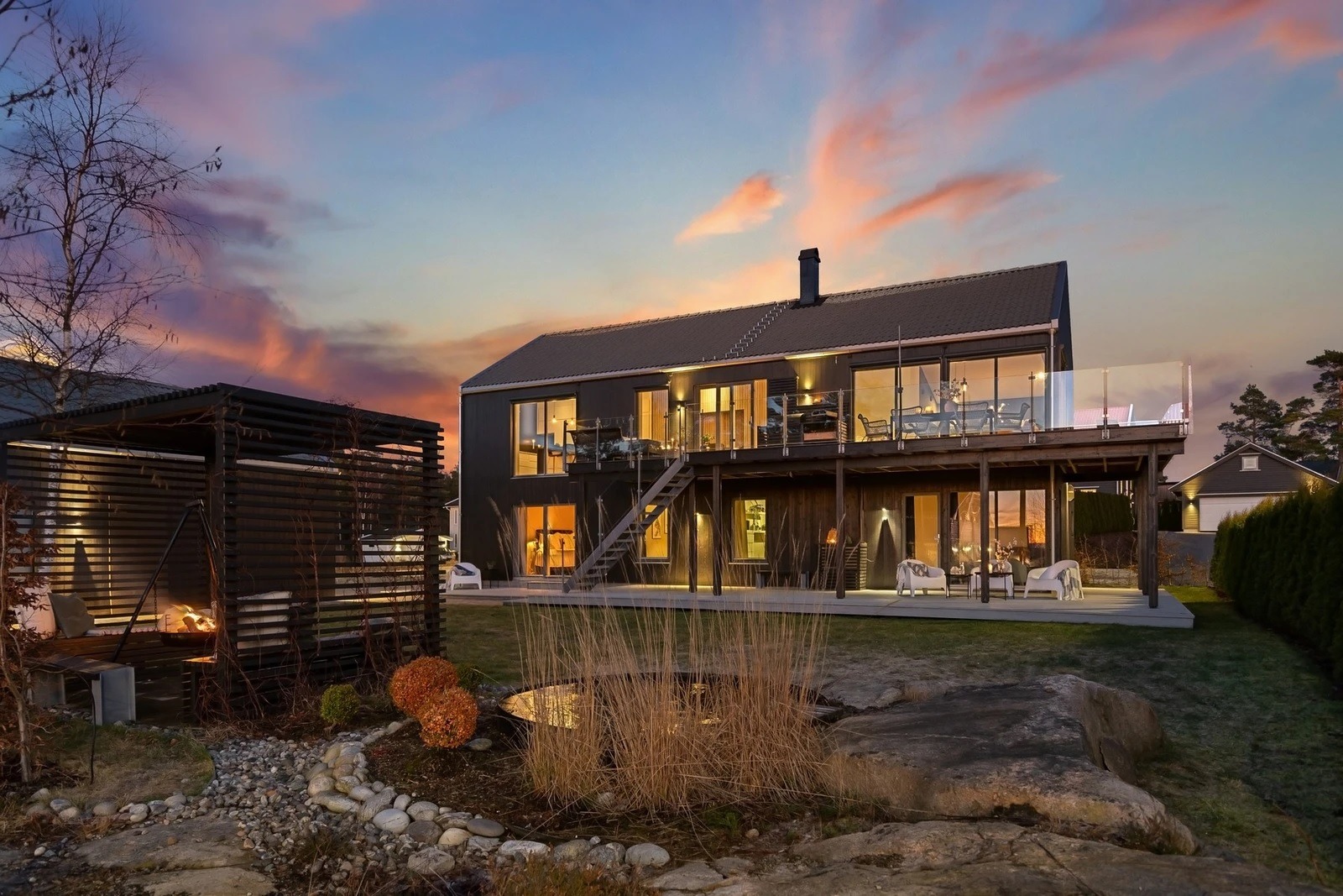 Dwukondygnacyjny dom z ciemną elewacją i dużym tarasem, widok z ogrodu z paleniskiem i oczkiem wodnym o zmierzchu.
