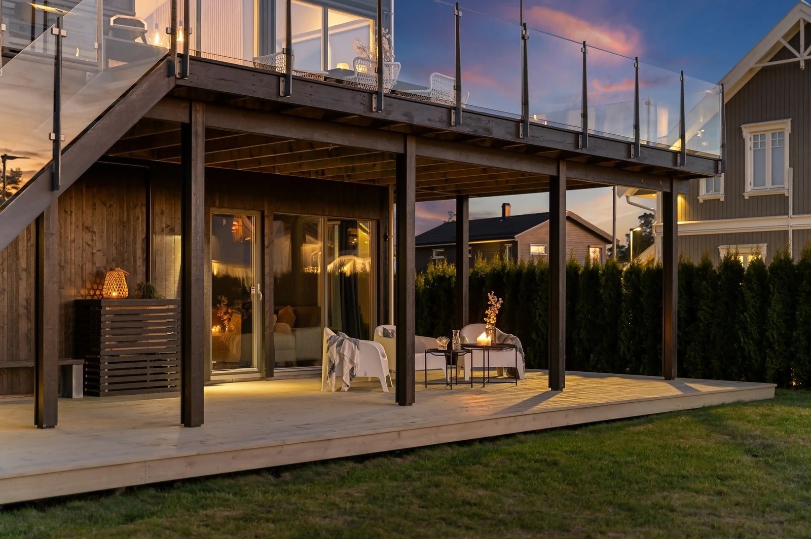 Dwupoziomowy taras przy domu z drewna, z przeszkloną balustradą i meblami ogrodowymi, oświetlony ciepłym światłem o zmierzchu.