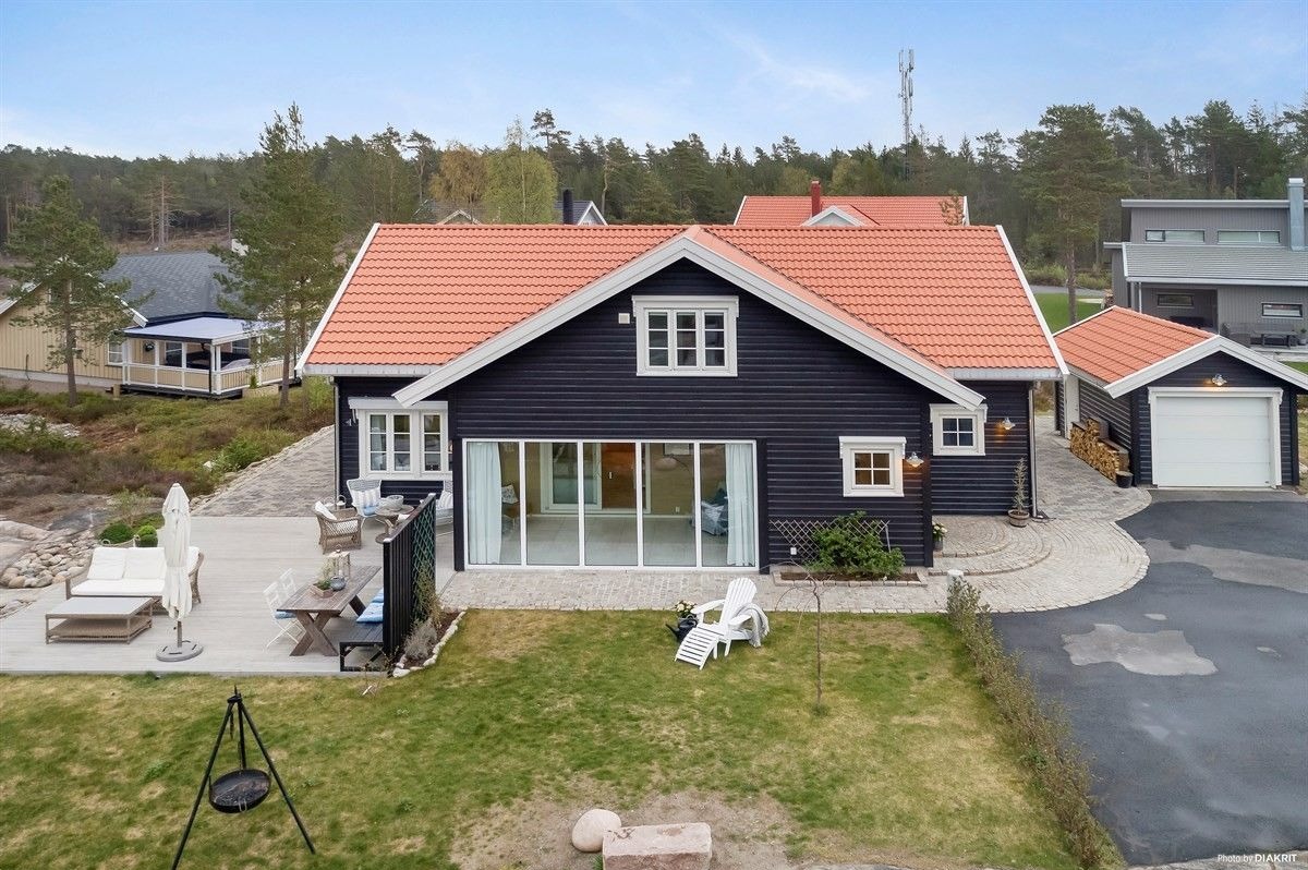 Czarny dom z pomarańczową dachówką i przeszkloną ścianą frontową, widok z góry na taras z meblami ogrodowymi i trawnik z grillem trójnożnym.