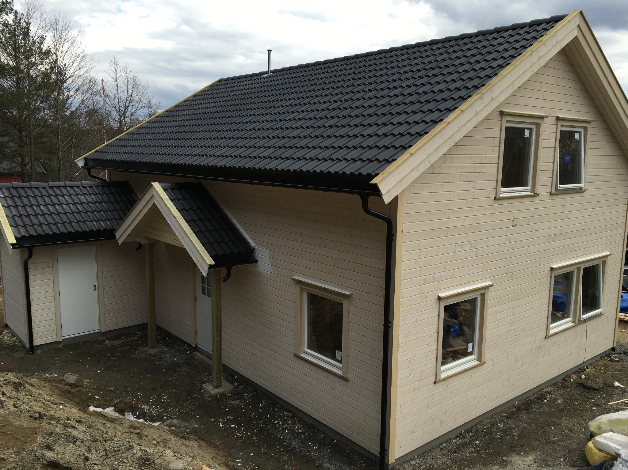 Surowy, jasny drewniany dom z ciemnym dachem z dachówki, widoczny z zewnątrz, z niezagospodarowanym terenem.