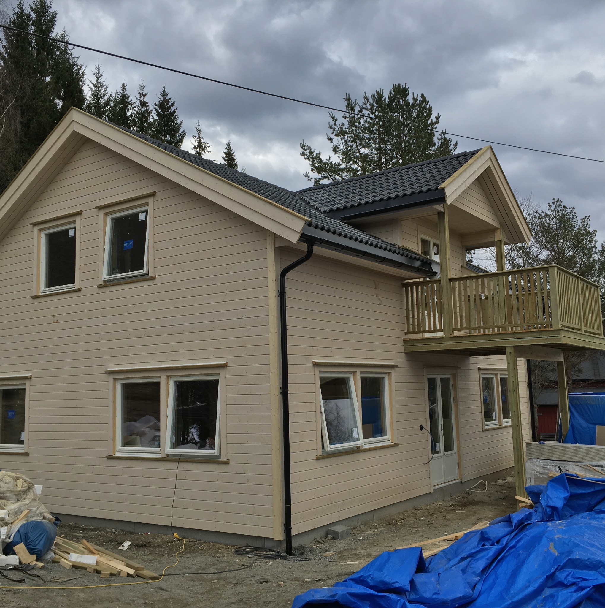 Surowy dom szkieletowy z drewnianym balkonem, widoczne nieotynkowane ściany, czarna rynna i niebieska plandeka budowlana na ziemi.