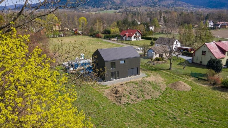 Nowoczesny, dwukondygnacyjny dom o elewacji w kolorze antracytu, z garażem wbudowanym w bryłę, usytuowany na lekko wzniesionej działce z widokiem na okoliczne zabudowania i wzgórza, sfotografowany...