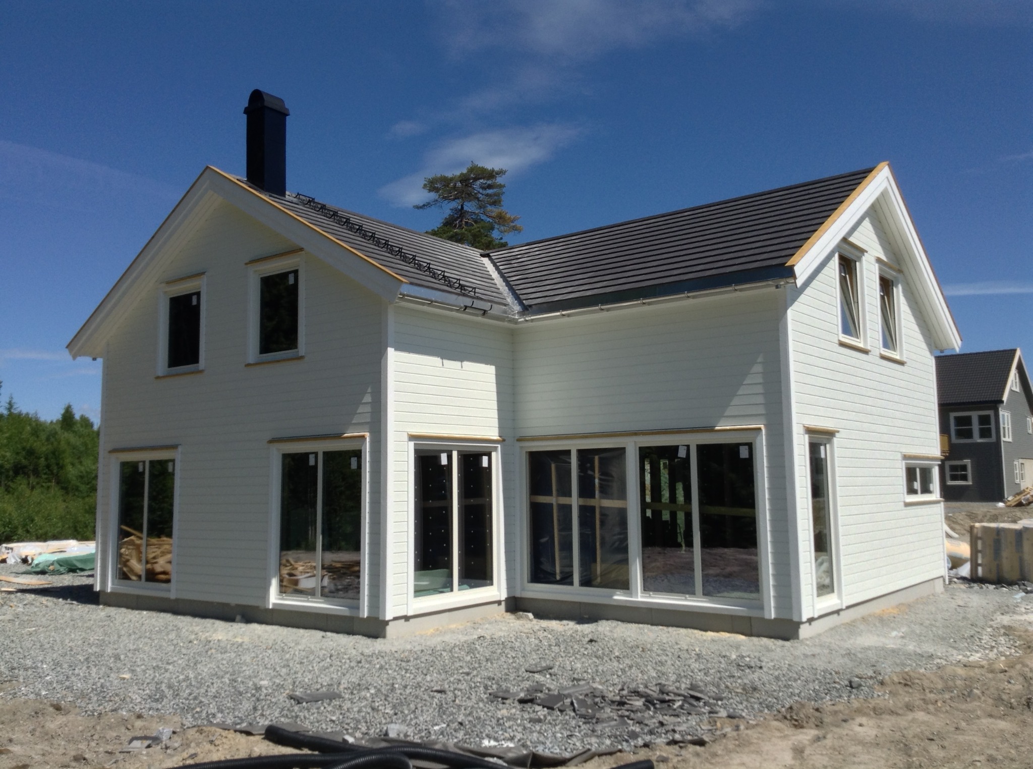 Biały, dwukondygnacyjny dom w trakcie budowy z czarnym kominem i szarą dachówką, widoczne okna bez szyb, teren wokół domu wysypany żwirem.