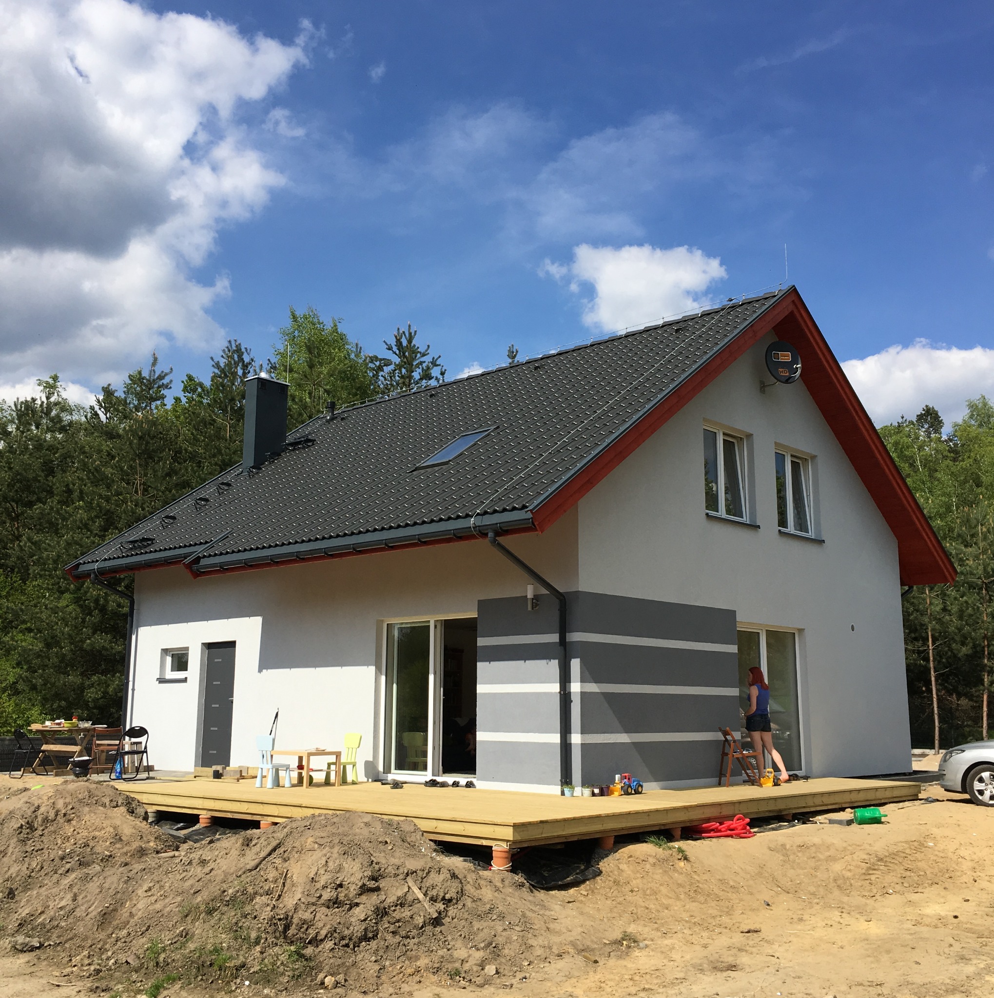 Nowoczesny dom jednorodzinny z szarą dachówką, drewnianym tarasem na podwyższeniu i elementami dekoracyjnymi w postaci szarych pasów na elewacji, w tle las i błękitne niebo z chmurami.