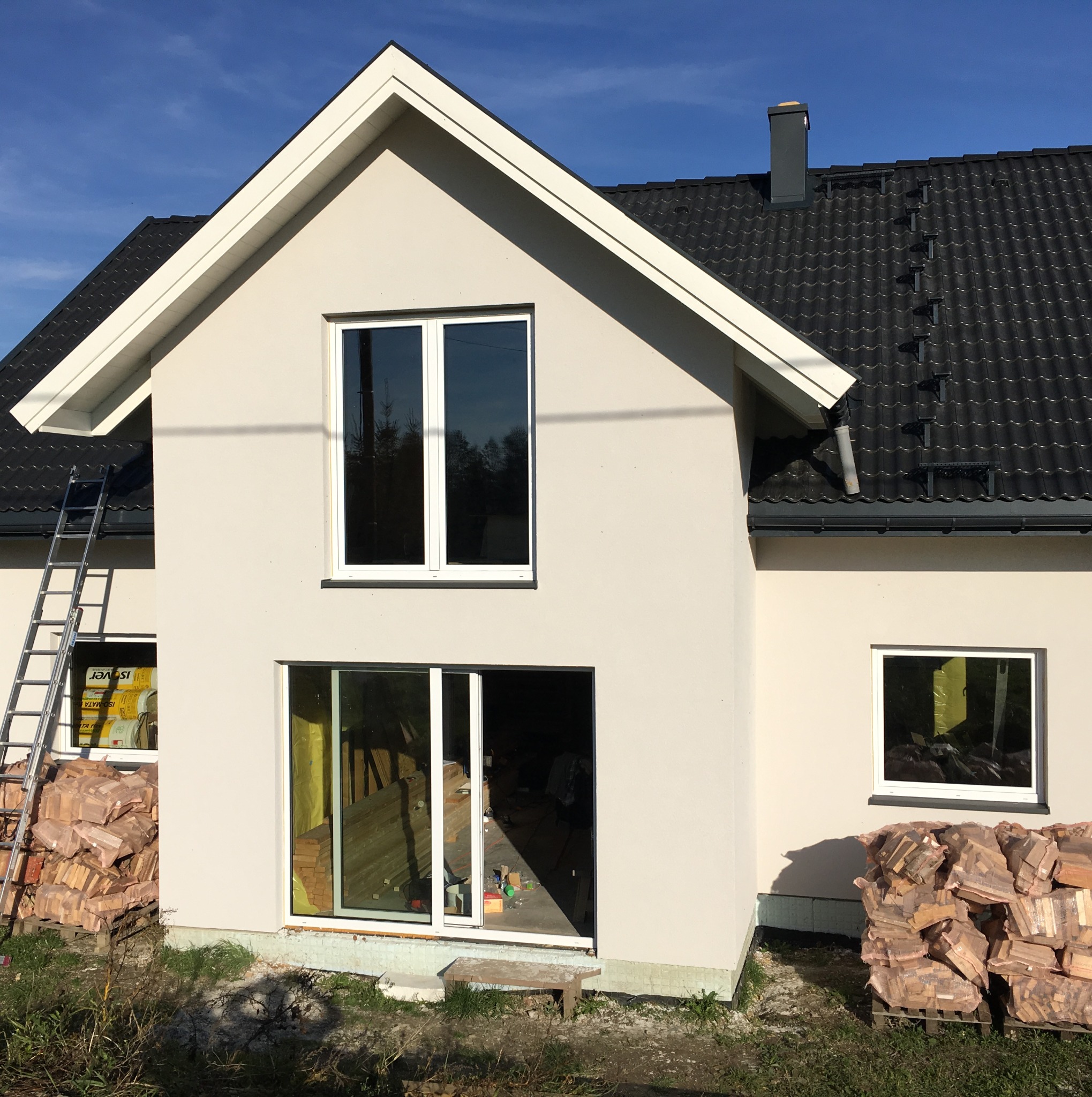 Dwukondygnacyjny dom w trakcie budowy z jasną elewacją, czarną dachówką i stosem drewna opałowego przy ścianie, z widoczną drabiną i oknami.