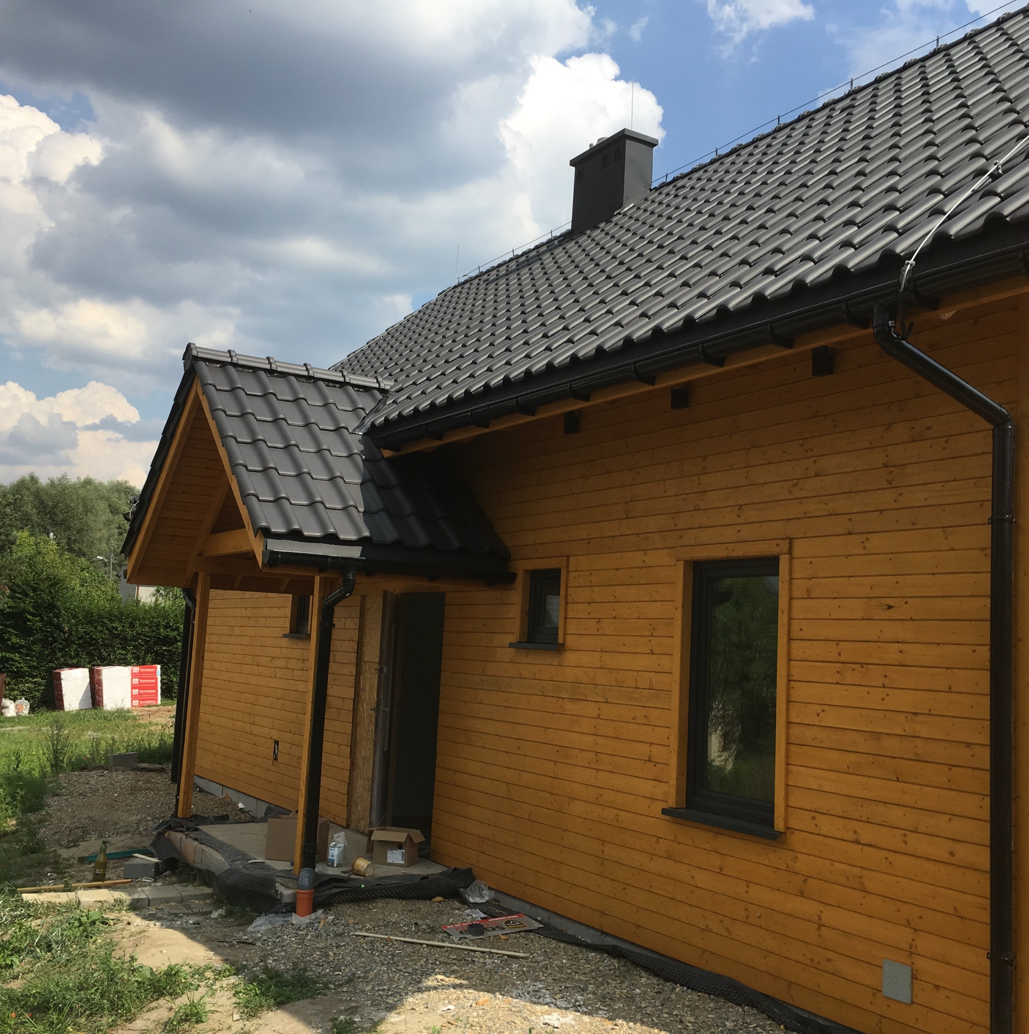Żółta elewacja domu w trakcie budowy, widoczny fragment dachu z czarną dachówką, okna z czarną stolarką, przygotowane materiały budowlane.