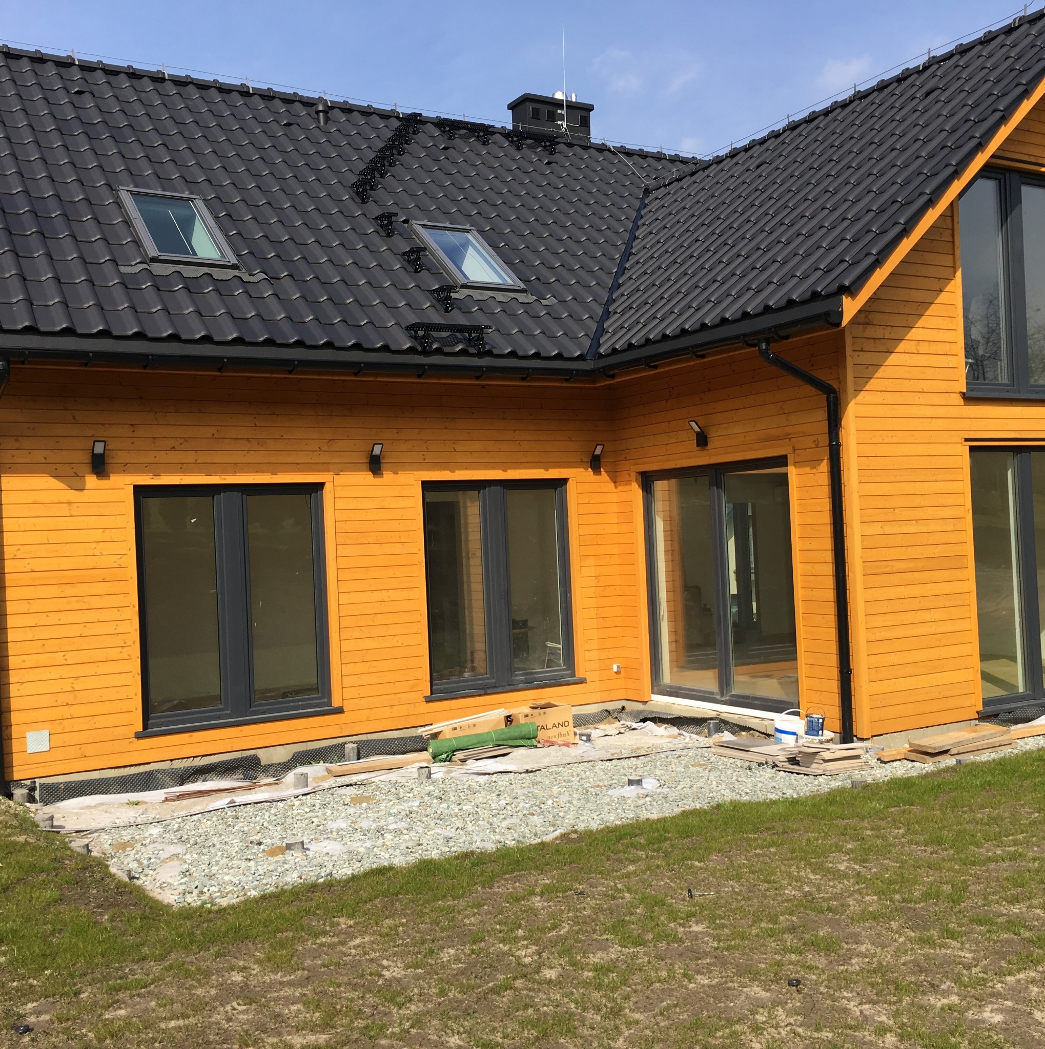 Żółta elewacja domu z drewna, czarny dach z oknami dachowymi, widoczne elementy przygotowania podłoża pod taras.
