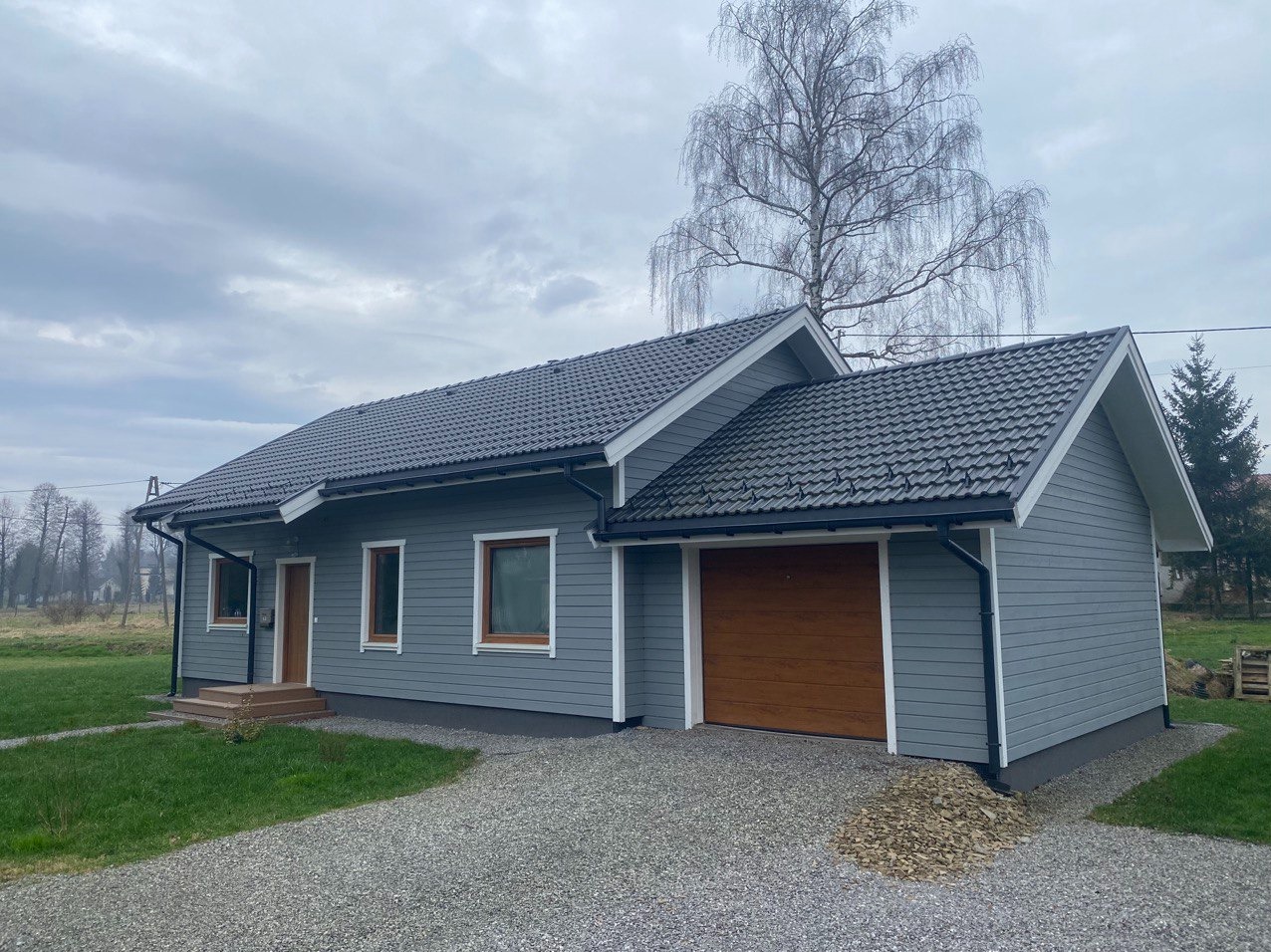 Gotowy, parterowy dom z szarą elewacją, brązowymi oknami i garażem z bramą segmentową, widok z zewnątrz na tle drzew i pochmurnego nieba.