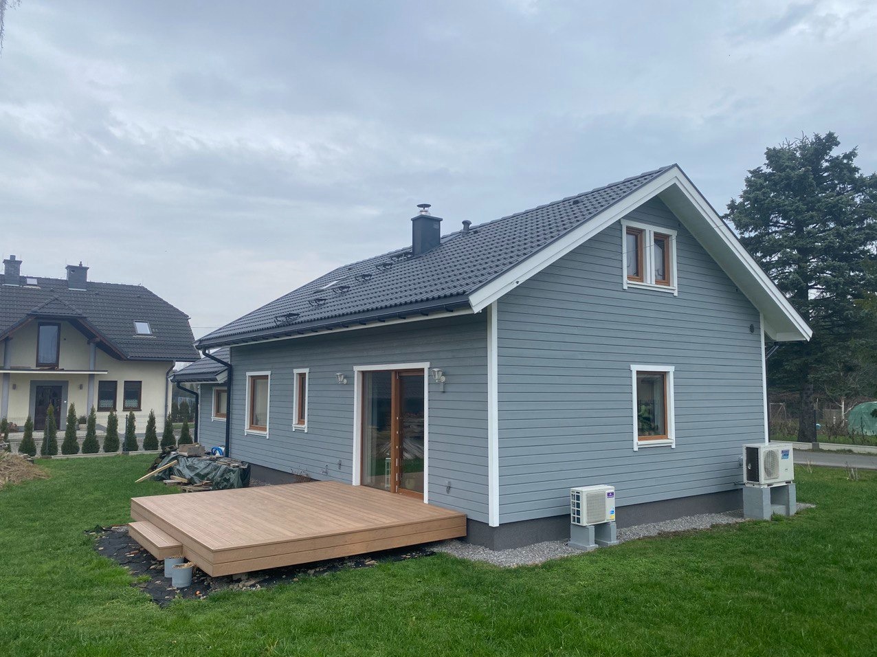 Szary dom z drewnianą elewacją i tarasem w trakcie budowy, widoczne klimatyzatory na zewnątrz.