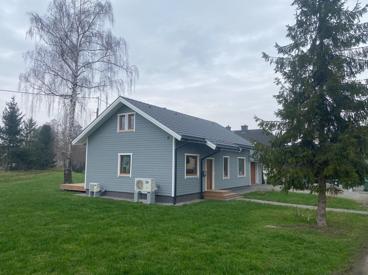 Gotowy, parterowy budynek z szarej deski z ciemnoszarą dachówką, dwa klimatyzatory na betonowych podstawach, drewniany taras, trawnik i drzewa na pochmurnym tle.
