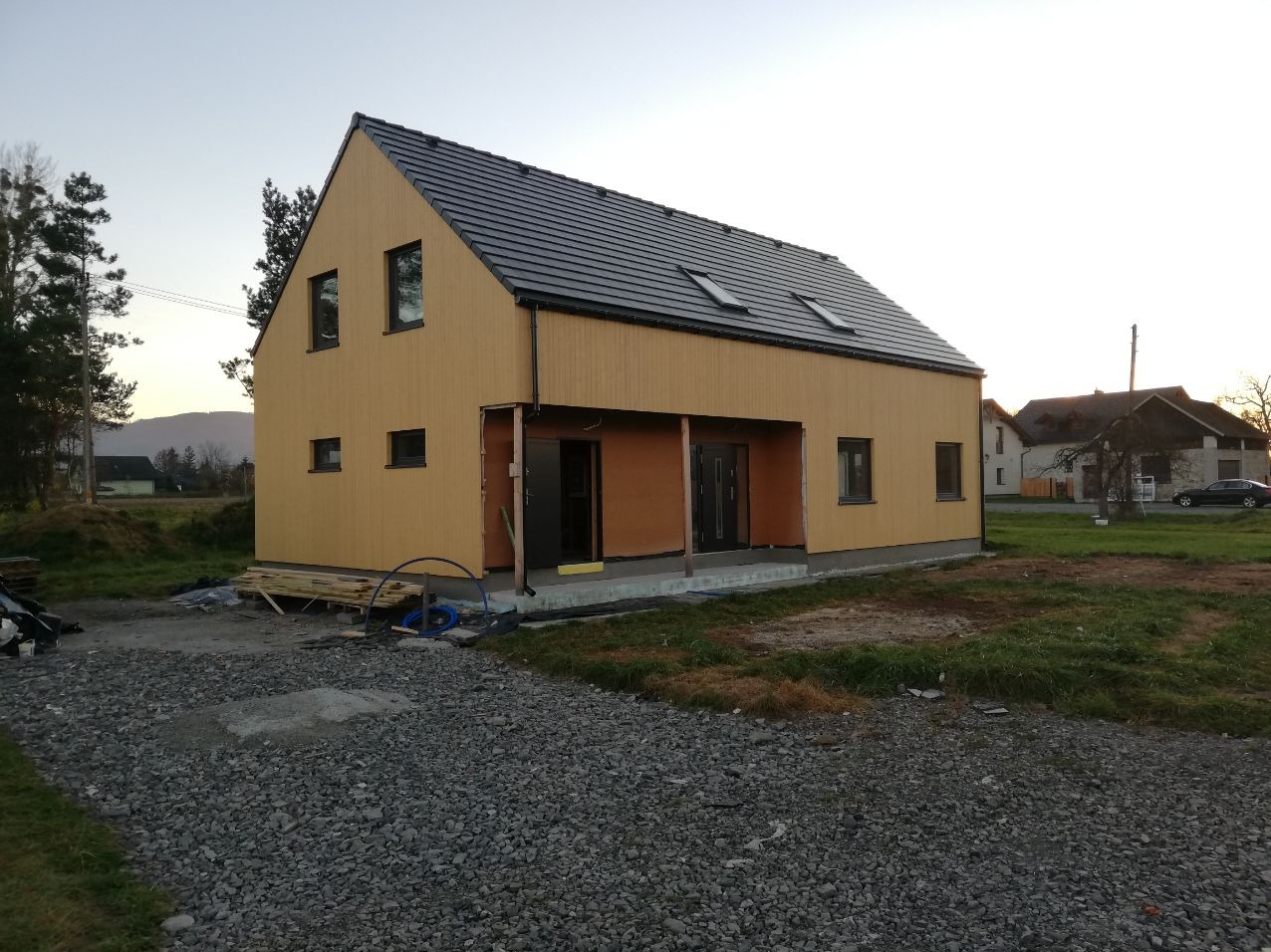 Nowoczesny, dwukondygnacyjny dom z drewnianą elewacją w odcieniach żółci, z ciemną dachówką i dwoma oknami dachowymi, w trakcie budowy, widoczne materiały budowlane i teren przygotowany pod ogród.