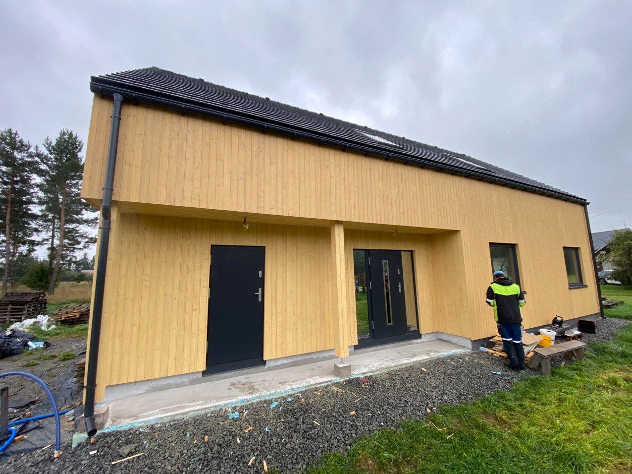 Nowoczesny, parterowy budynek z jasną, drewnianą elewacją pionową, czarnym dachem i stolarką okienną w kolorze antracytowym, na tle pochmurnego nieba, z pracownikiem w odblaskowej kamizelce przy...