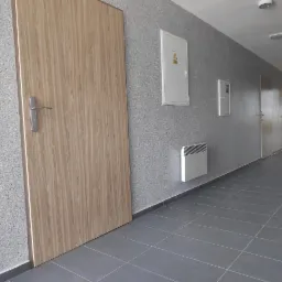 Korytarz biurowy z drewnopodobnymi drzwiami, szarą, chropowatą ścianą i szarą podłogą z płytek; widoczne skrzynki elektryczne i grzejnik na ścianie.
