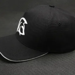 Czarna czapka z daszkiem z białym haftowanym logo przedstawiającym stylizowaną literę lub symbol graficzny, leżąca na ciemnoszarym tle o wyraźnej fakturze.