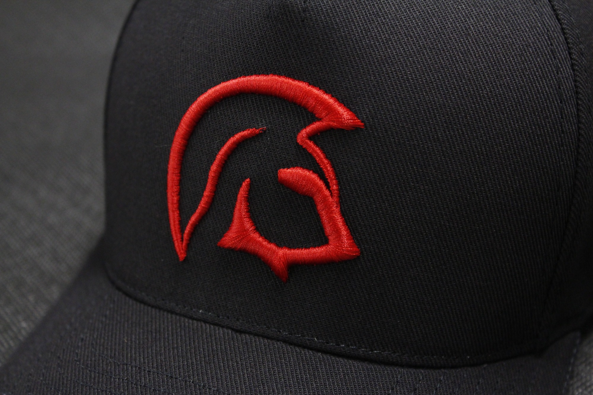 Czarna czapka z daszkiem z wyhaftowanym czerwonym logo przedstawiającym stylizowany hełm.