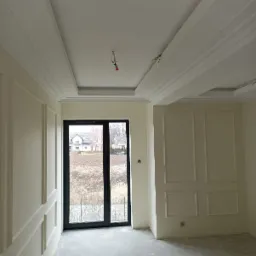 Pokój z wykończonymi ścianami i sufitem w stylu klasycznym, listwy dekoracyjne, duże okno z widokiem na domy w tle, surowy beton na podłodze, przygotowany do dalszych prac wykończeniowych.
