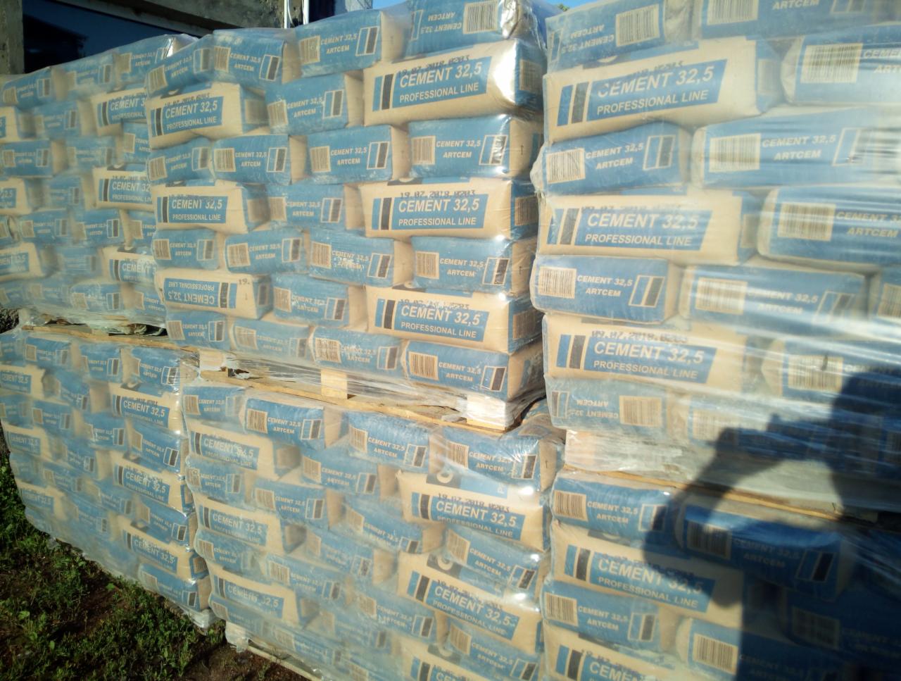 Paleta worków z cementem 32,5, owinięta folią stretch, składowana na zewnątrz, widoczne cienie i fragment zieleni u dołu.