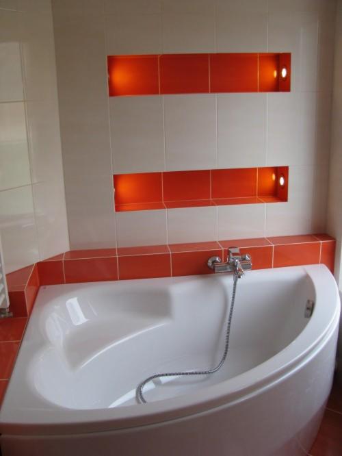 Biała wanna narożna z chromowaną baterią i wężem prysznicowym na tle ściany wyłożonej białymi płytkami z dwoma poziomymi, pomarańczowymi wnękami z oświetleniem.