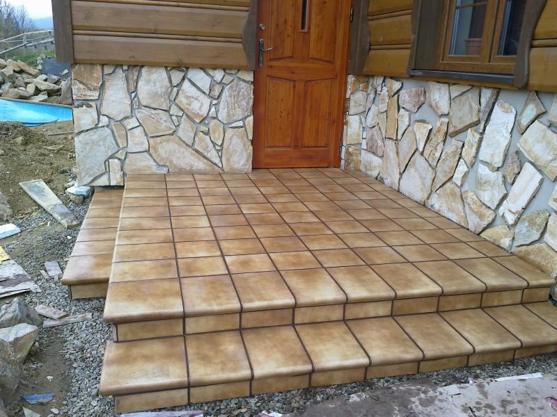 Wyłożone brązowymi płytkami schody wejściowe do drewnianego domu z kamienną elewacją, widoczne fugi i faktura płytek.