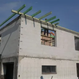 Wznoszenie ścian domu z bloczków betonowych, widoczne zielone belki stropowe i pracownik w oknie, etap budowy w słoneczny dzień.