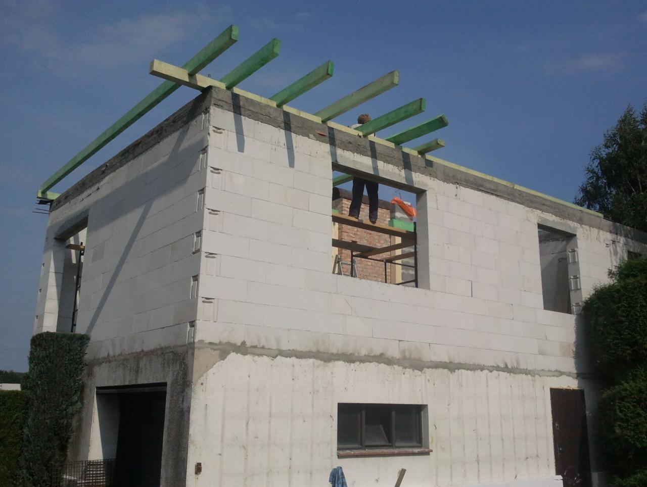 Wznoszenie ścian domu z bloczków betonowych, widoczne zielone belki stropowe i pracownik w oknie, etap budowy w słoneczny dzień.