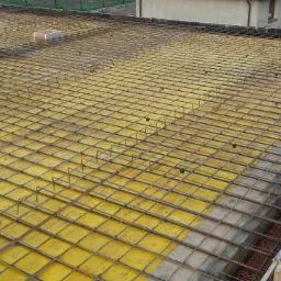 Zbrojenie stropu z siatki stalowej na żółtym deskowaniu, widoczne elementy murowane ścian i przygotowane szalunki.