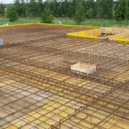 Zbrojenie płyty fundamentowej z żółtym szalunkiem na obrzeżach i drewnianymi elementami szalunkowymi w tle, widok z góry na rozległy teren budowy.