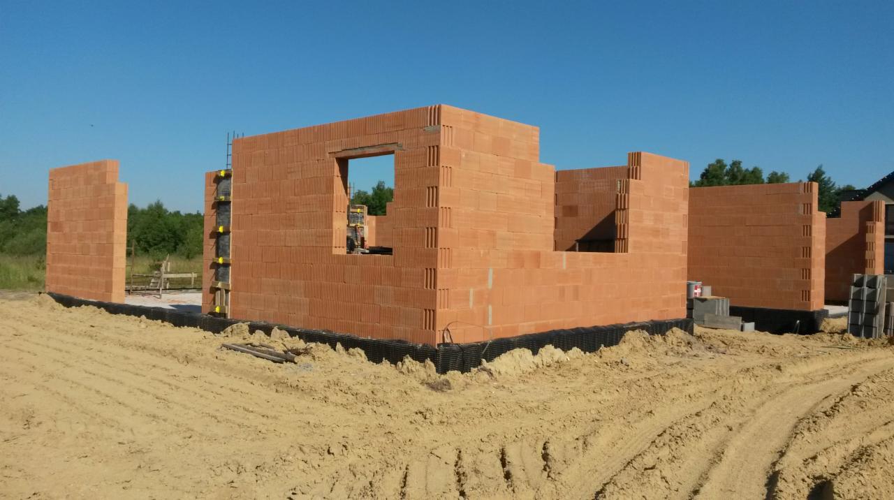 Wznoszenie ścian parteru z bloczków ceramicznych na fundamencie, widoczne zabezpieczenie przeciwwilgociowe, teren budowy w słoneczny dzień.