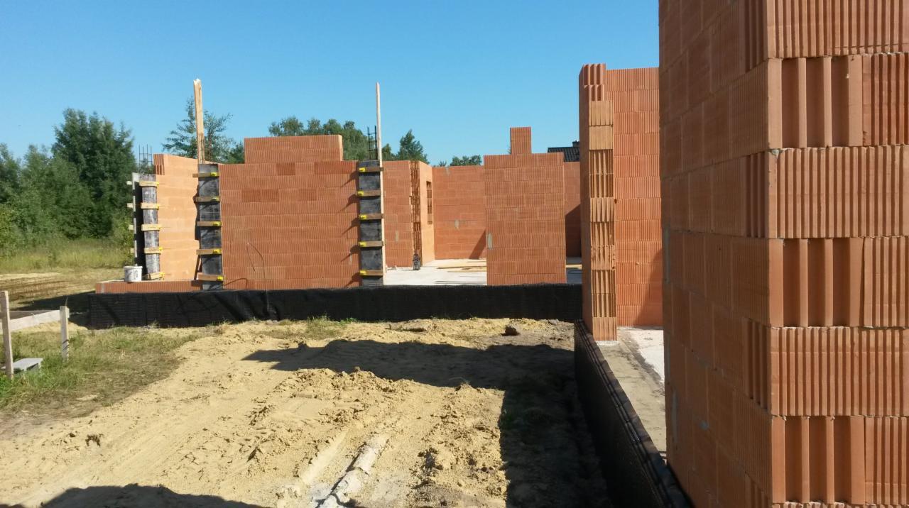 Budowa domu z czerwonej cegły ceramicznej, widoczne ściany w trakcie wznoszenia, szalunki fundamentowe z czarną folią hydroizolacyjną, teren budowy z piaskiem i trawą.