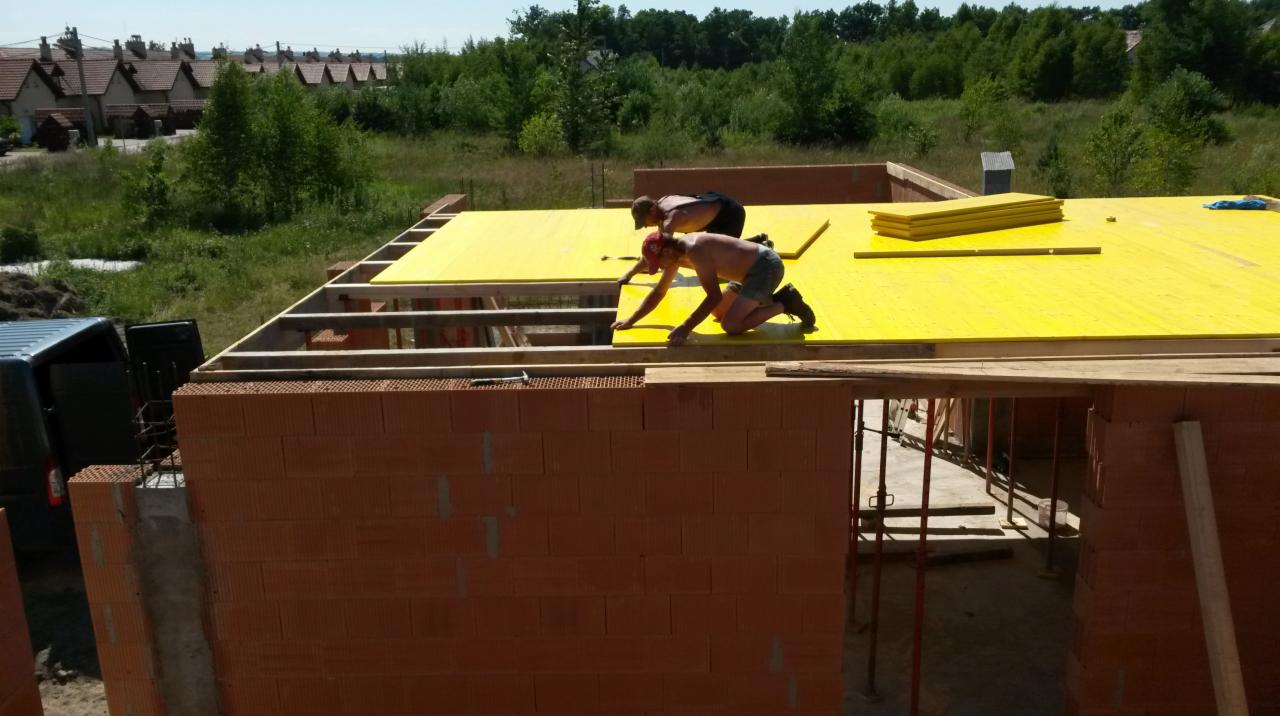 Dwóch robotników bez koszulek układa żółte płyty na dachu budowanego domu z czerwonej cegły, w tle zabudowa jednorodzinna i zieleń.