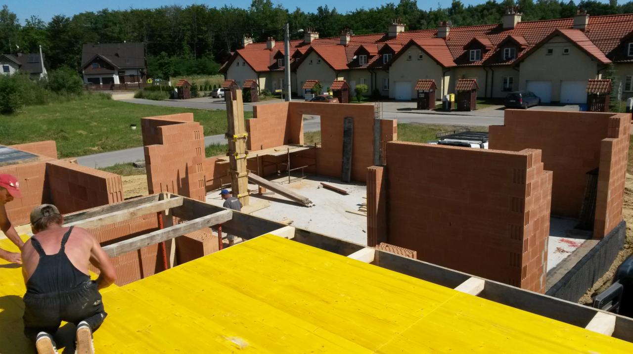 Budowa domu z czerwonej cegły ceramicznej, widoczne wznoszone ściany na fundamencie, żółte szalunki stropowe, w tle zabudowa szeregowa.