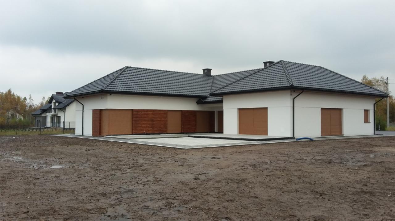 Nowoczesny, parterowy dom z szarą dachówką i brązowymi roletami zewnętrznymi, widok z zewnątrz na częściowo zagospodarowaną działkę w pochmurny dzień.