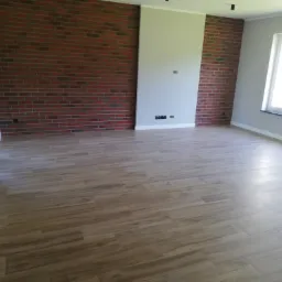 Jasne, przestronne wnętrze z drewnopodobną podłogą, fragmentem ściany z cegły klinkierowej i świeżo pomalowaną ścianą w kolorze szarym, widoczne gniazdka elektryczne i okno.