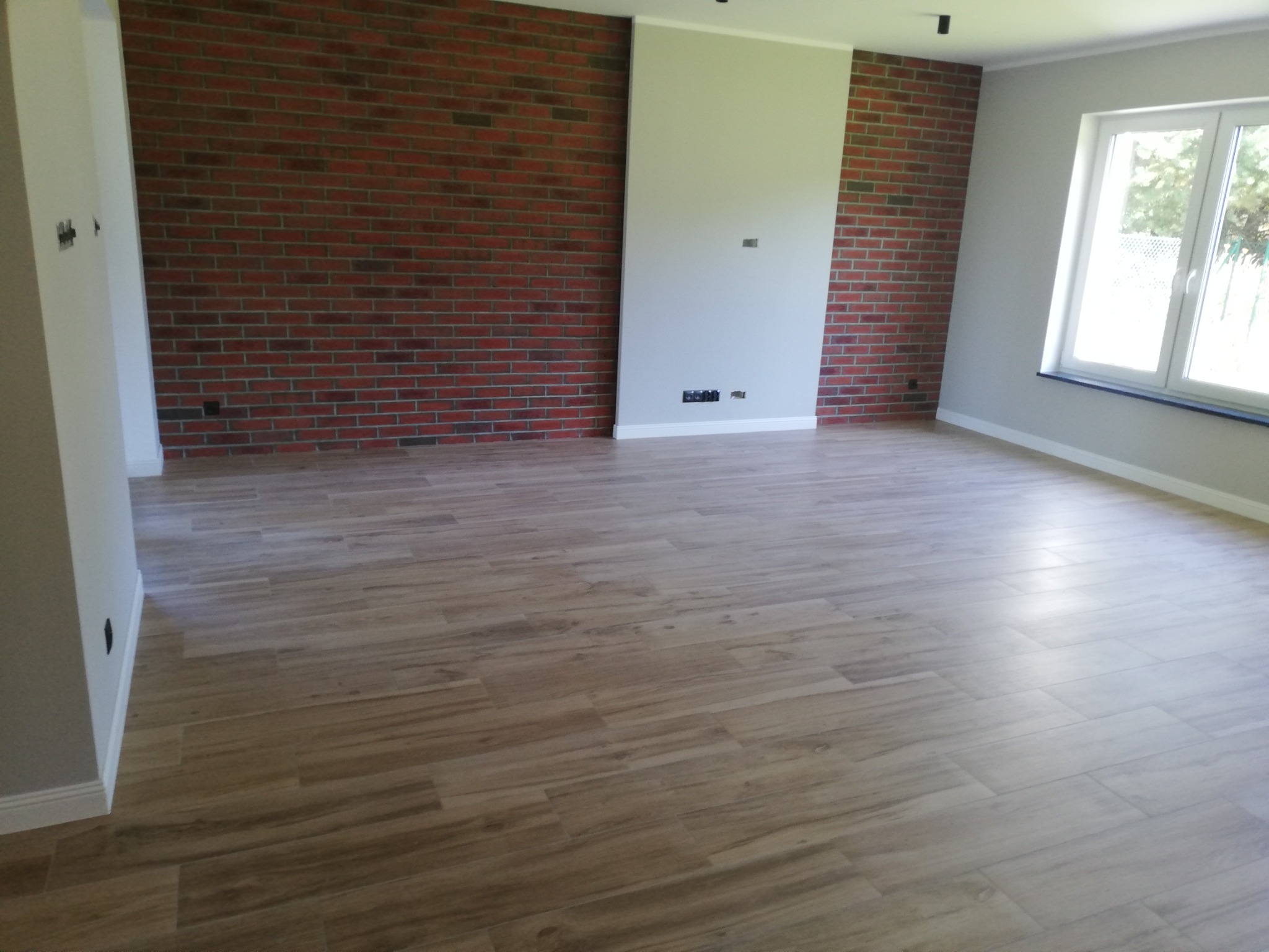 Jasne, przestronne wnętrze z drewnopodobną podłogą, fragmentem ściany z cegły klinkierowej i świeżo pomalowaną ścianą w kolorze szarym, widoczne gniazdka elektryczne i okno.