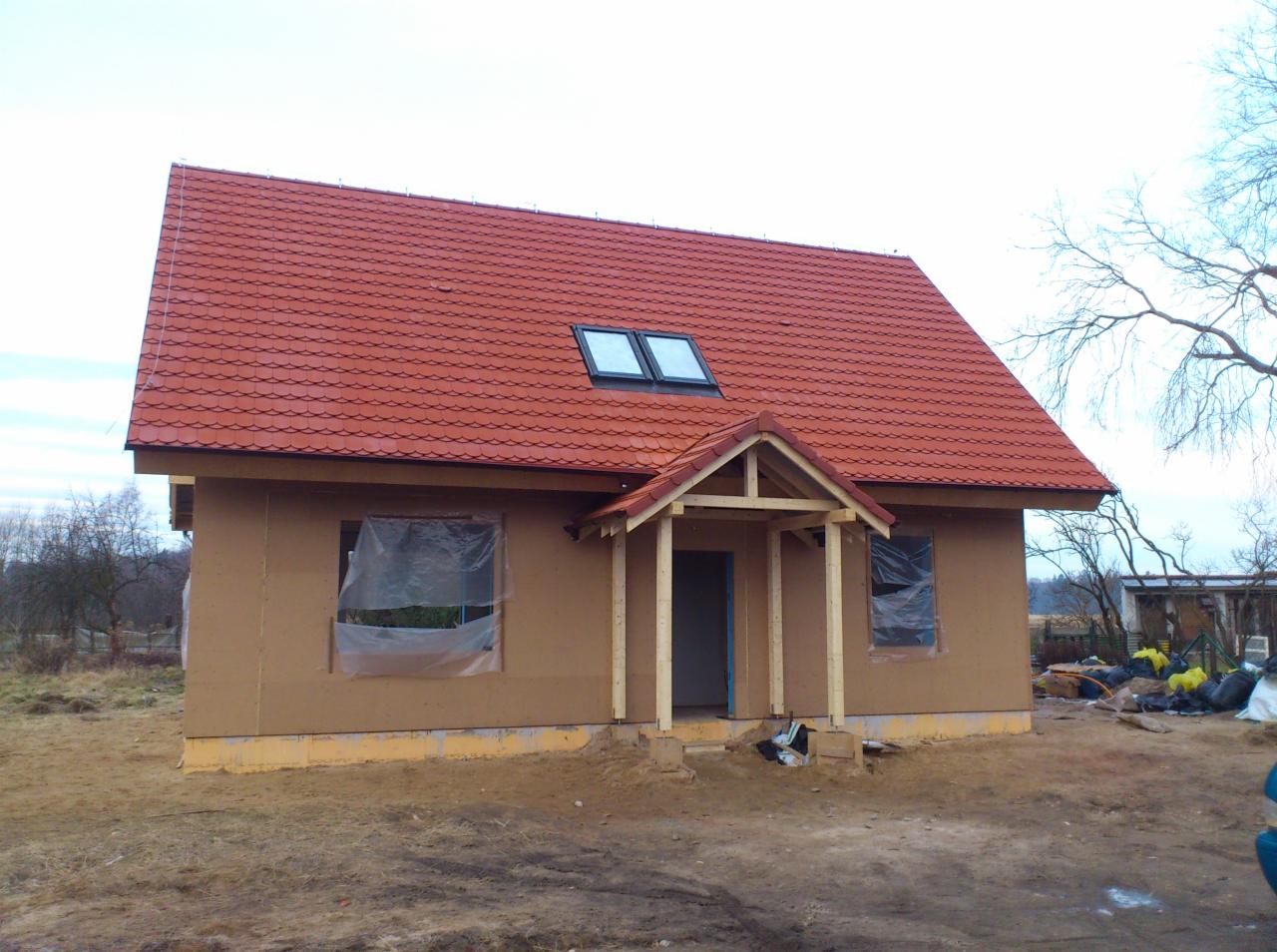 Dom w trakcie budowy z nowym, czerwonym dachem ceramicznym i oknami dachowymi, widoczne zabezpieczone folią okna i drewniana konstrukcja ganku.