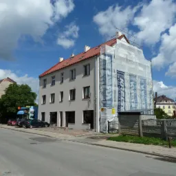 Budynek wielorodzinny w trakcie prac elewacyjnych, osłonięty siatką z reklamą producenta materiałów budowlanych, widok z ulicy w słoneczny dzień.