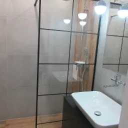 Nowoczesna łazienka z kabiną prysznicową typu walk-in z czarnymi profilami, białą umywalką nablatową i drewnianymi akcentami. Minimalistyczny design i jasne oświetlenie.