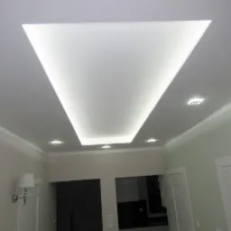 Biały sufit podwieszany z trapezowym wgłębieniem oświetlonym taśmą LED, uzupełniony o listwy przysufitowe i punktowe lampy halogenowe w jasnym wnętrzu.