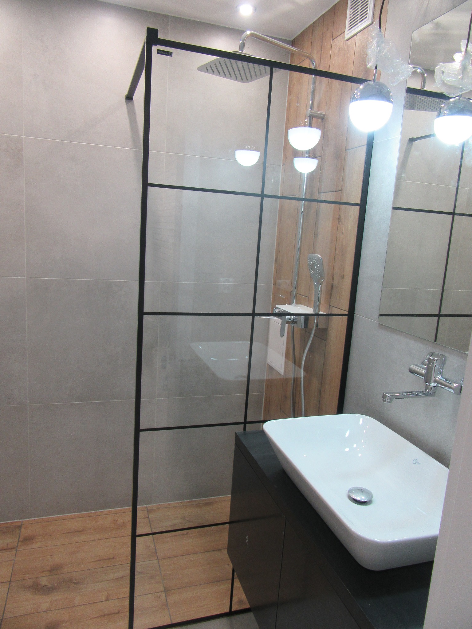 Nowoczesna łazienka z kabiną prysznicową typu walk-in z czarnymi profilami, białą umywalką nablatową i drewnianymi akcentami. Minimalistyczny design i jasne oświetlenie.