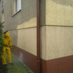 Skala - Pracownik w żółtym kombinezonie czyści elewację budynku za pomocą myjki ciśnieniowej. Widoczny kontrast między czystą a brudną powierzchnią ściany.