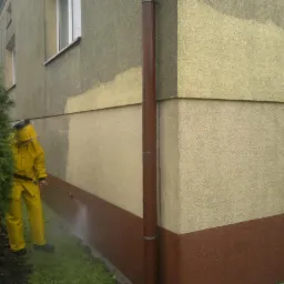 Pracownik w żółtym kombinezonie czyści elewację budynku za pomocą myjki ciśnieniowej. Widoczny kontrast między czystą a brudną powierzchnią ściany.