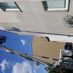 Skala - Pracownik w koszu podnośnika czyści elewację budynku. Widoczne fragmenty umytej i nieumytej powierzchni, ukazujące efekt pracy. Błękitne niebo w tle.