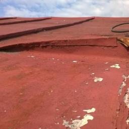 Skala - Czerwony dach z blachy z łuszczącą się farbą, widoczny przewód i fragment okna dachowego.