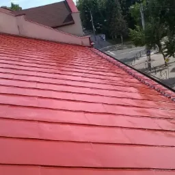 Czerwony, błyszczący dach z blachy dachówkowej po czyszczeniu, widok z góry. W tle budynki, drzewa i ulica z zaparkowanymi samochodami, słoneczny dzień.