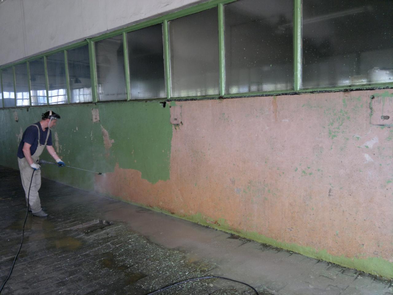 Pracownik w kombinezonie czyści ścianę budynku przemysłowego za pomocą myjki ciśnieniowej, widoczne okna i fragment posadzki. Częściowo usunięta warstwa farby.