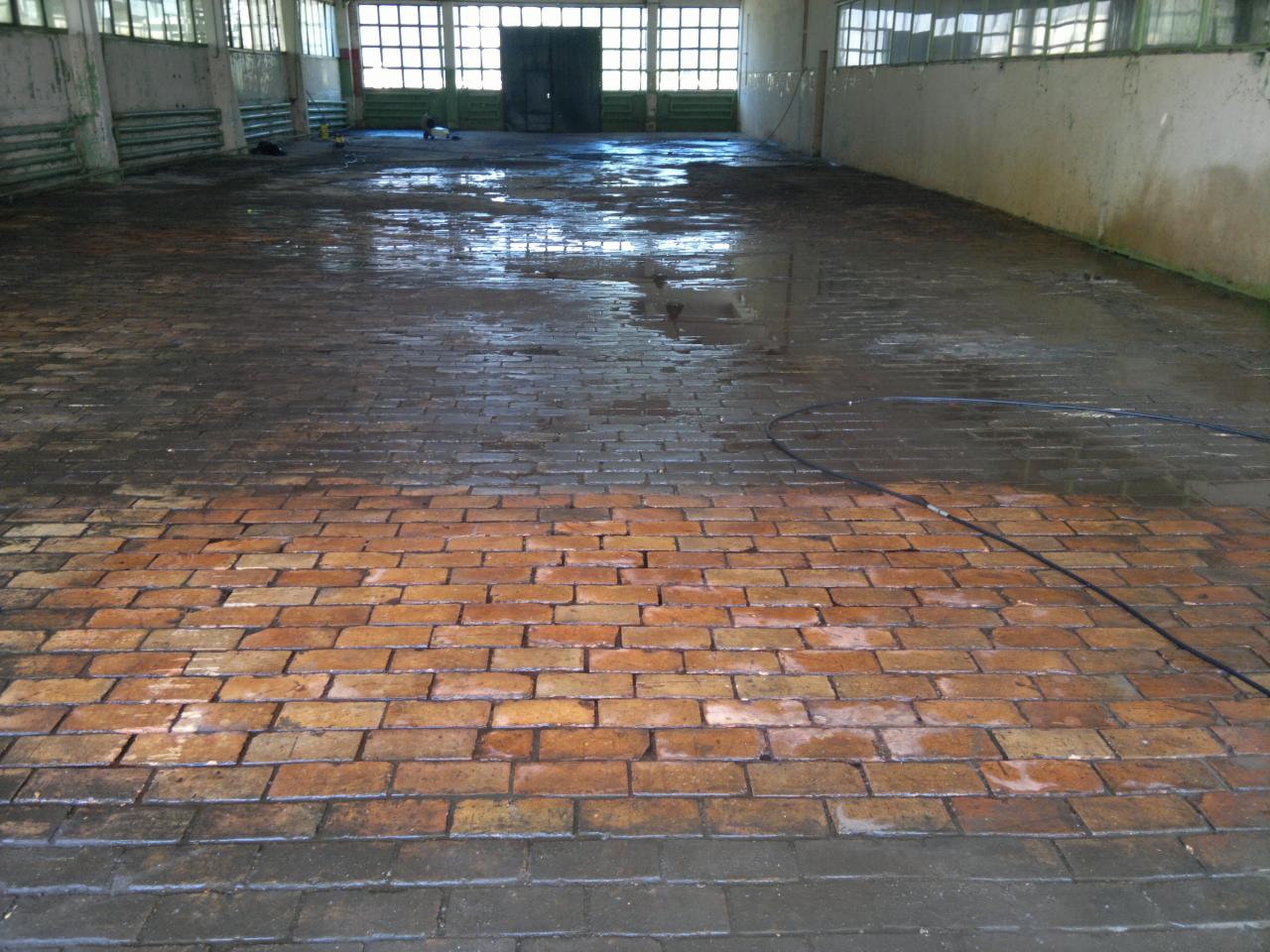 Częściowo umyta, mokra, ceglana posadzka w hali przemysłowej, z widocznym kablem i refleksami światła na powierzchni. Kontrast czystej i brudnej powierzchni.