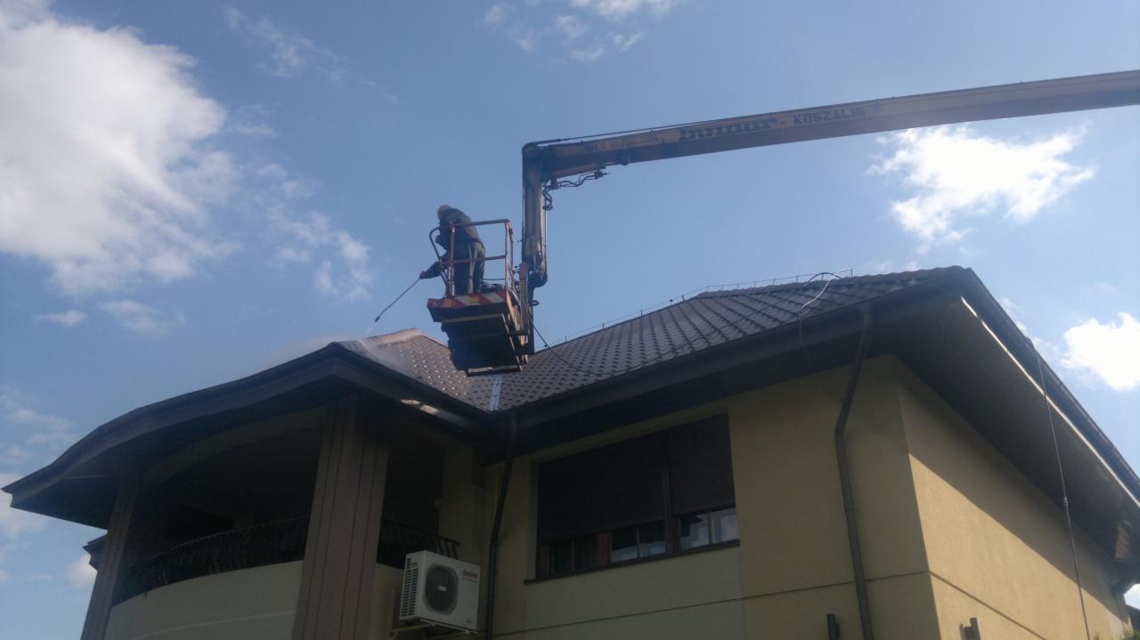 Pracownik na podnośniku czyści dach domu jednorodzinnego pod ciśnieniem. Widoczna instalacja klimatyzacji na ścianie budynku i błękitne niebo z chmurami w tle.