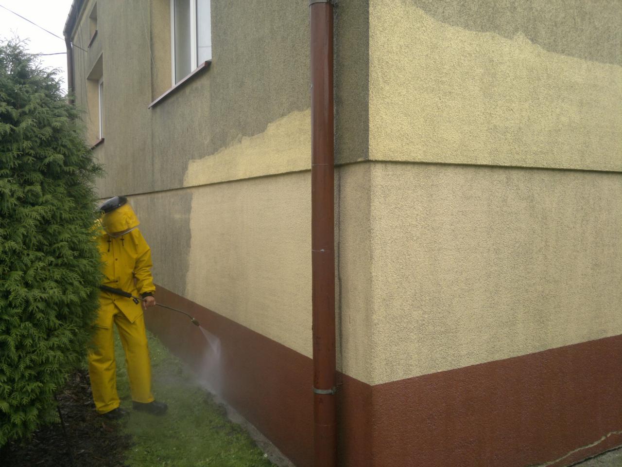 Pracownik w żółtym kombinezonie czyści elewację budynku za pomocą myjki ciśnieniowej. Widoczny kontrast między czystą a brudną powierzchnią ściany.