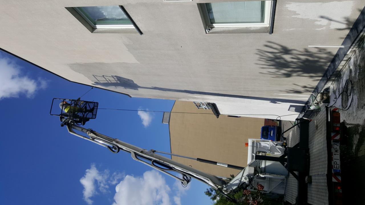 Pracownik w koszu podnośnika czyści elewację budynku. Widoczne fragmenty umytej i nieumytej powierzchni, ukazujące efekt pracy. Błękitne niebo w tle.