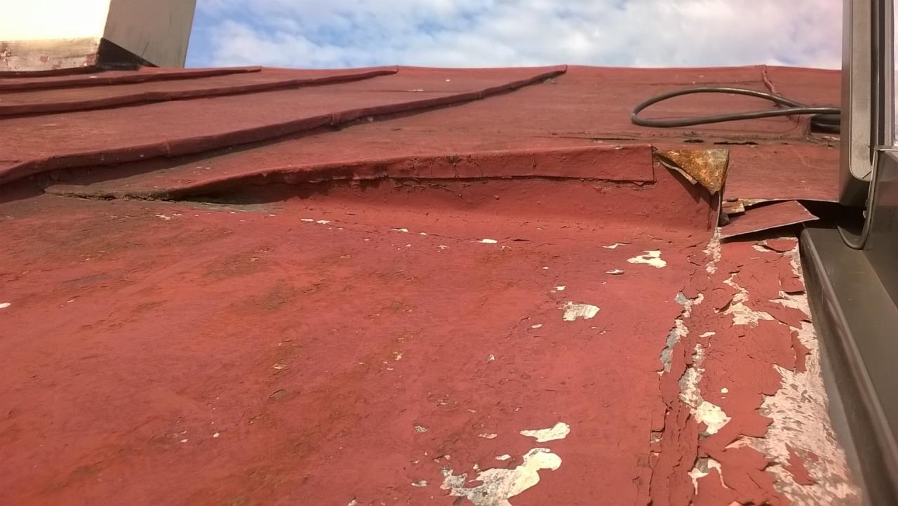 Czerwony dach z blachy z łuszczącą się farbą, widoczny przewód i fragment okna dachowego.